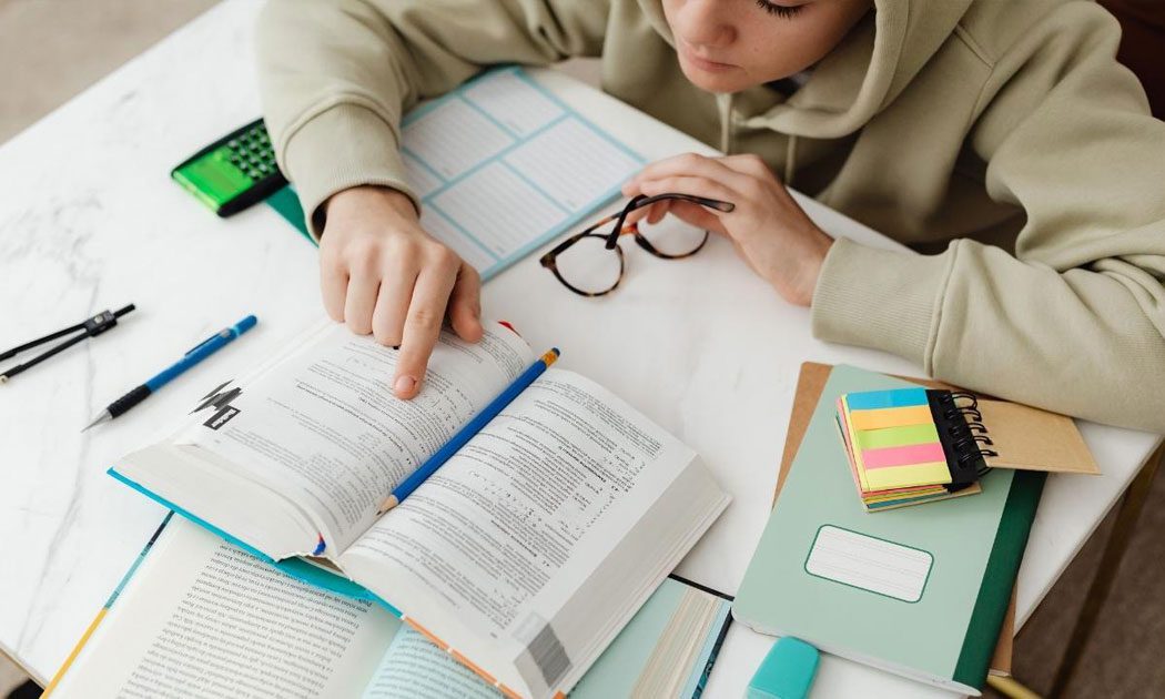 Student Studying, Looking at a Book with Glasses in Hand, Surrounded by Books
